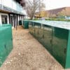 A row of large, green metal storage units, designated as e-bike storage lockers, line the side of a building along a gravel pathway. Another building and trees are visible in the background. The tops of the units are wet, suggesting recent rain.