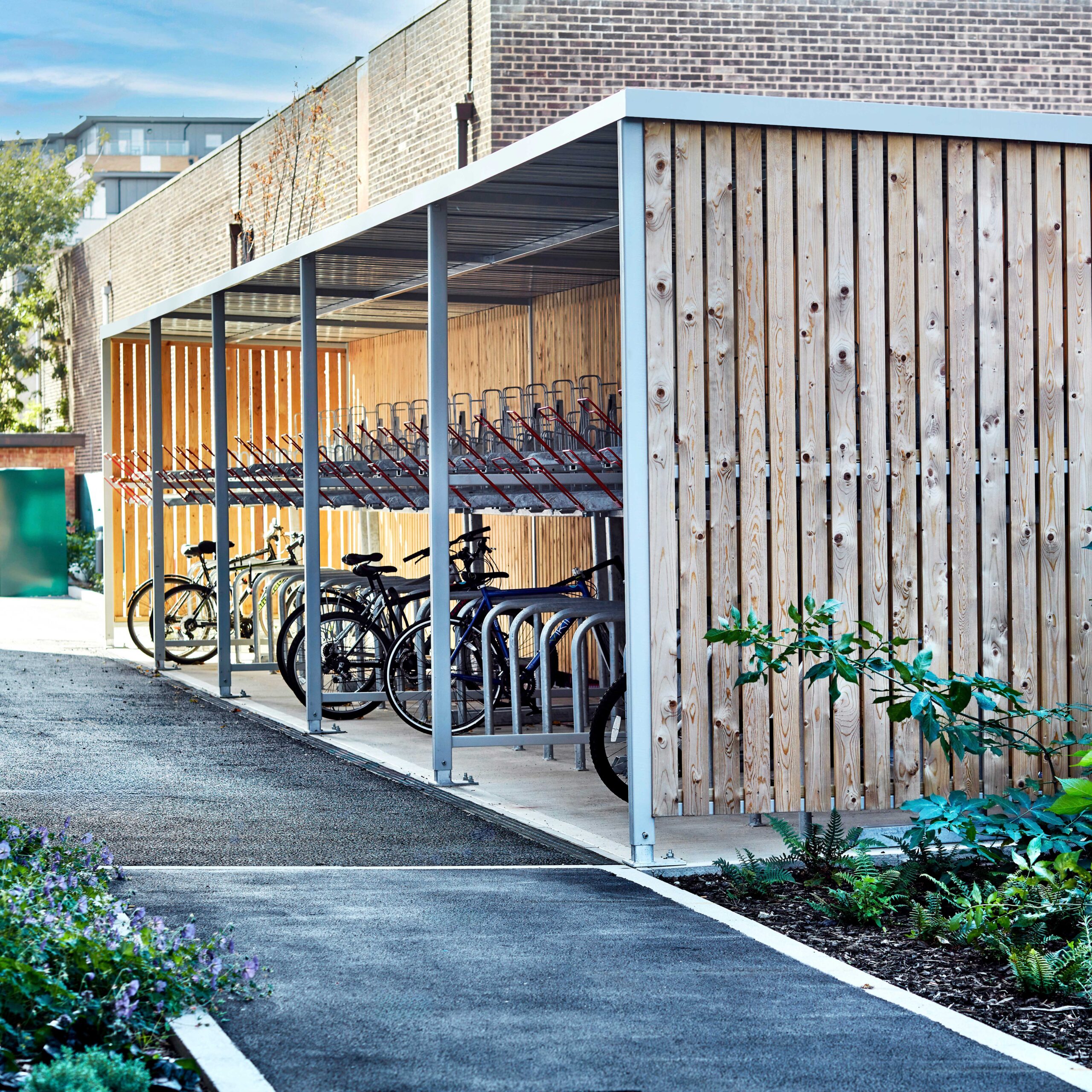 A modern, covered bicycle parking area with empty racks and a few parked bicycles, featuring a wooden back wall and metal supports.
