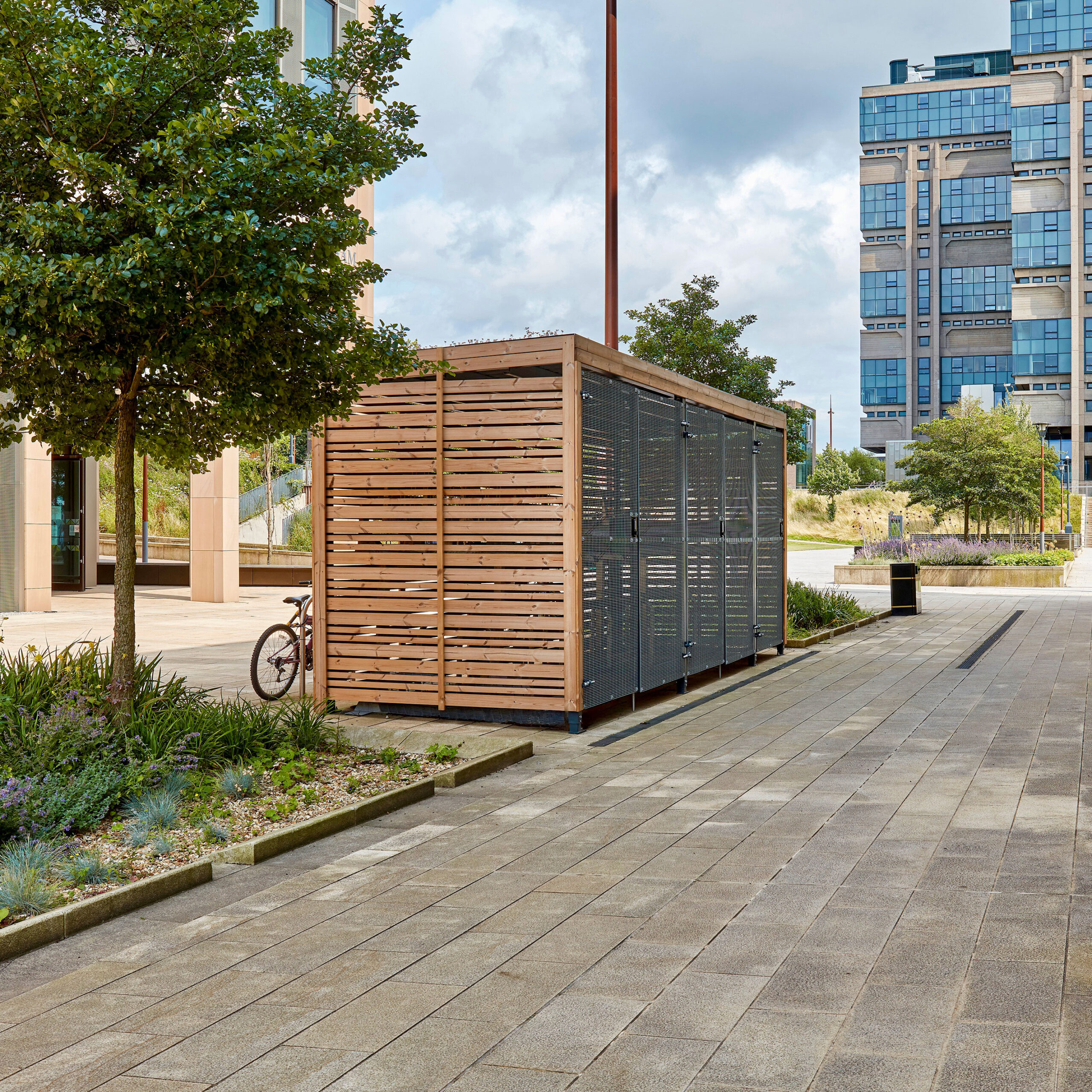A modern wooden bicycle storage shed with metal elements, located in an urban plaza with paved pathways, landscaping, and tall buildings in the background. a red bicycle is parked next to it.