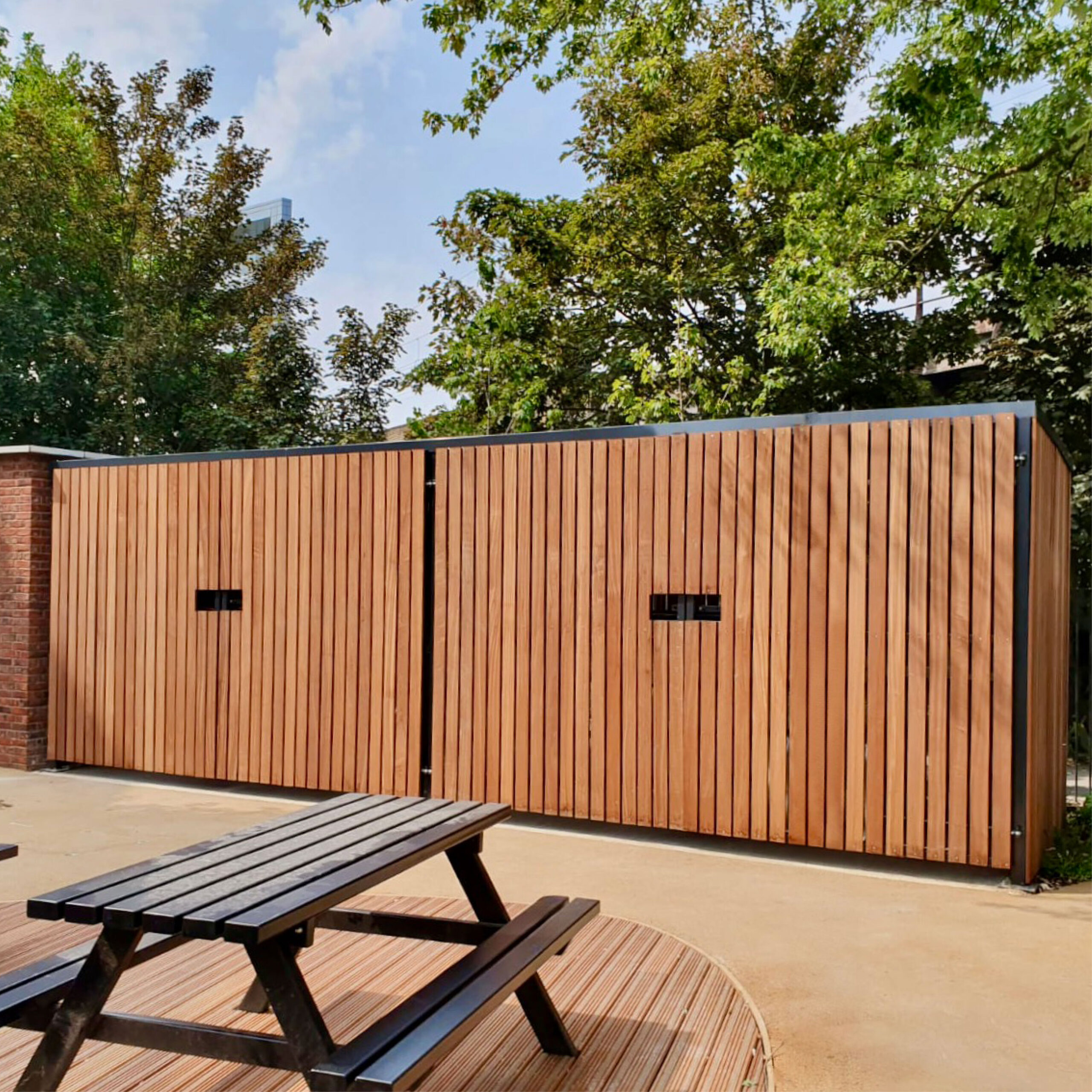 A two-tier wooden cycle shelter with closed swing doors underneath green trees