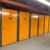 A row of storage units with bright orange doors and dark frames in an industrial setting. Each door has a number label, including 12 and 13 visible. Overhead pipes and fluorescent lighting can be seen in the concrete hallway—ideal for high-security bike storage in residential property development projects.