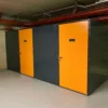 A dimly lit underground storage area with three metal storage units. Each unit, designed for high-security bike storage, has a bright orange door with a small black sign displaying the unit number. The surrounding walls and ceiling are unfinished, with visible pipes and concrete, ideal for a Tenant Cycle Store.