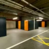 A row of brightly lit storage units in an indoor facility, perfect for storing bikes. Each unit has a door painted in orange with black trim, numbered 10 through 14, and is equipped with a lock. Overhead, there are exposed pipes and a long fluorescent light fixture—ideal for the Tenant Cycle Store's high-security space.