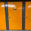 A row of bright orange storage lockers with dark gray frames is perfect for a Tenant Cycle Store, offering a high-security space. The middle locker has a metal sign displaying the number 13. All lockers have black handles and a smooth, shiny finish. The ceiling above has exposed red piping.
