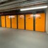 A high-security bike storage area is situated in a corridor with several bright orange doors, numbered, and set in grey frames. The ceiling has exposed pipes and ducts. The floor is clean and smooth, well-lit by overhead fluorescent lights. A directional sign is visible on the left wall.
