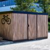 A modern wooden bike shed with a large bicycle symbol, located outside an office building in a sunny environment.