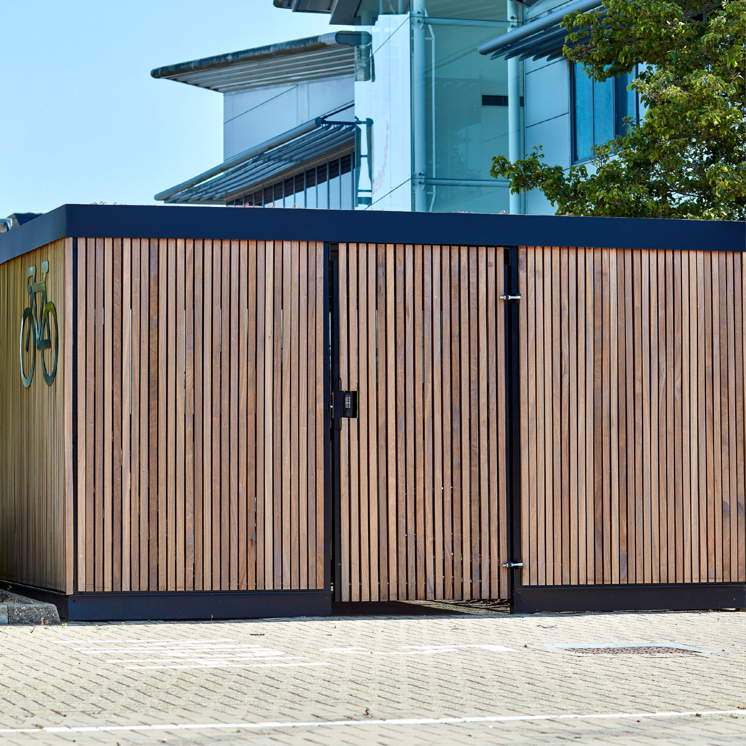A modern wooden bike storage facility with vertical slats, featuring a black bicycle symbol and double doors, located next to an urban building with a glass exterior.
