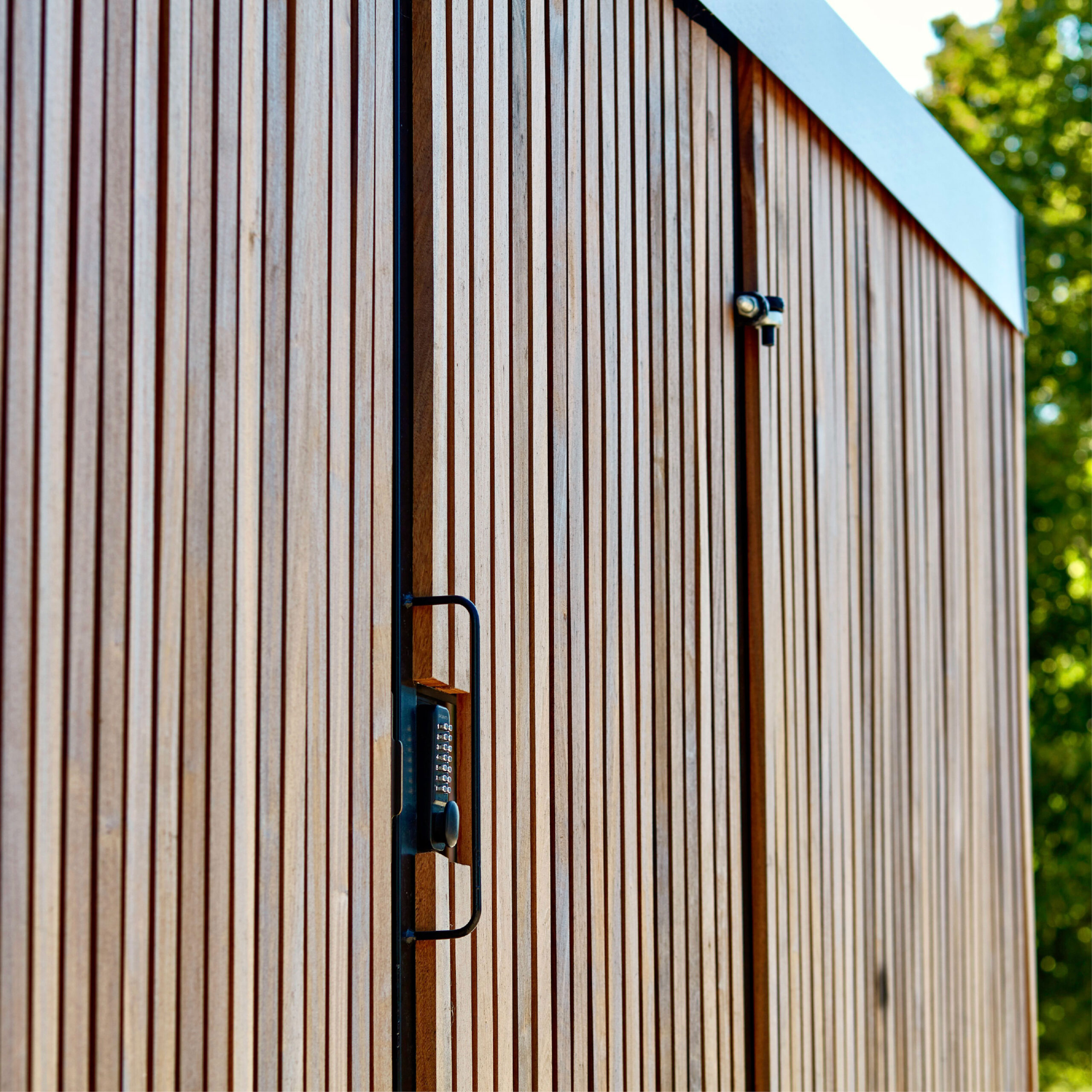 Vertical wooden slats form a wall with a sharp geometric metal handle on a door, highlighted by sunlight. a small security camera is mounted near the top of the frame.