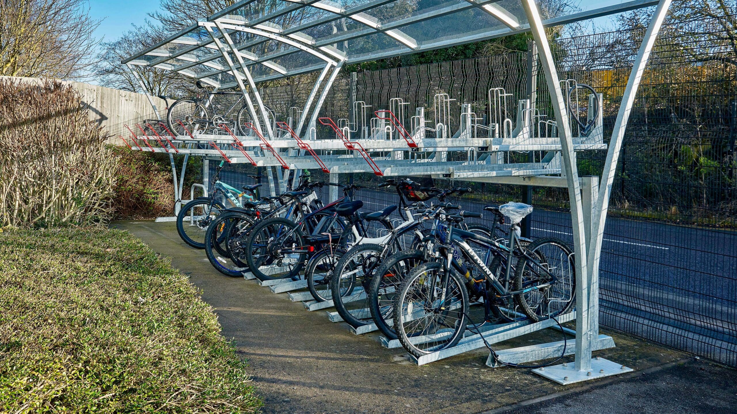 A modern, Two Tier bike rack covered by a Chelsea Cycle Shelter that is a transparent overhead shelter, with several bicycles parked in the lower section. The background features a hedge and trees, against a clear blue sky.