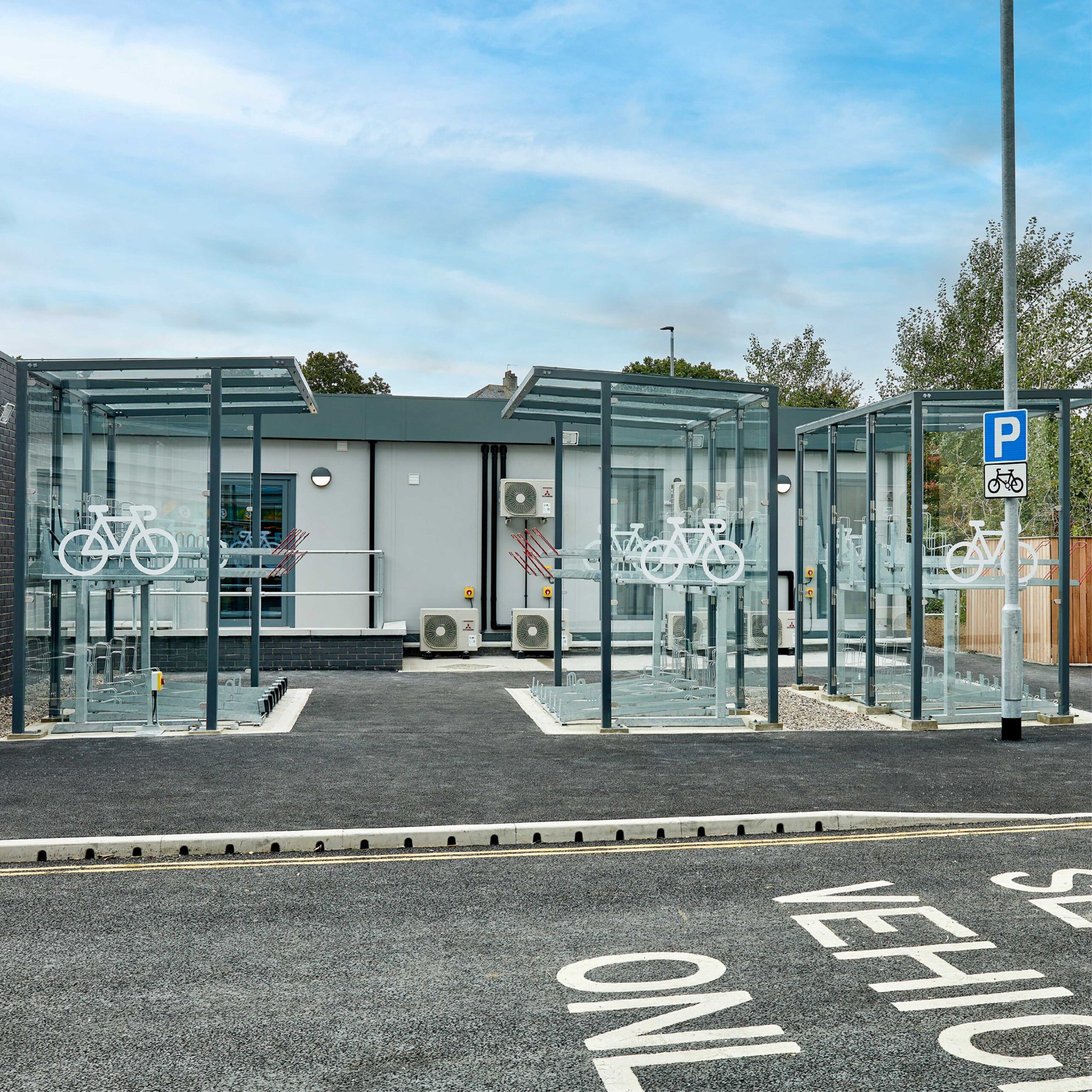 Modern bicycle parking facility with glass enclosures and clear bicycle icons on the entrances, located next to a parking area with white pavement markings indicating "shelter.