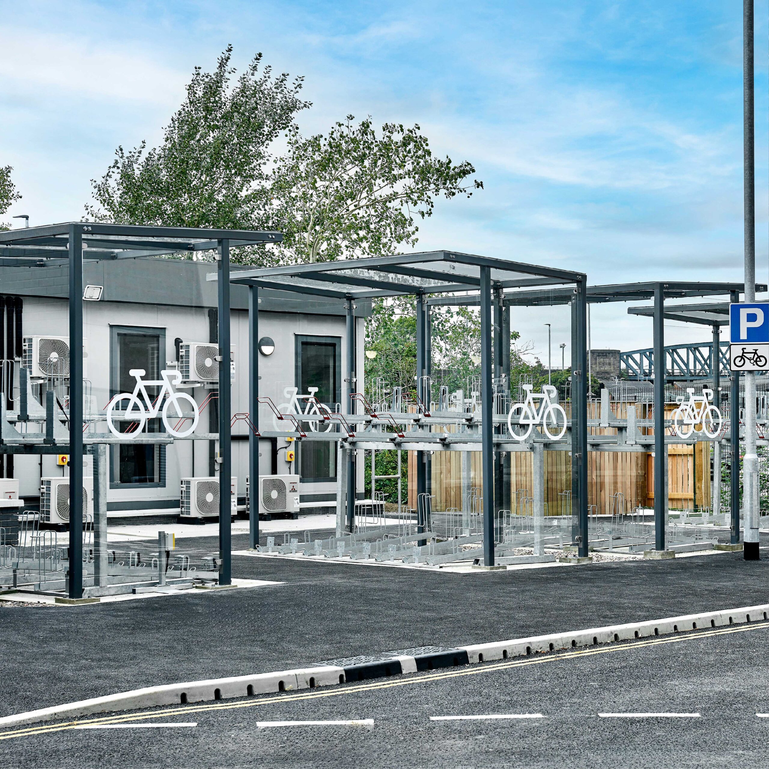 Modern bicycle parking facility with covered bike racks and secured bicycle stands beside a paved road and parking lot, under a clear sky.