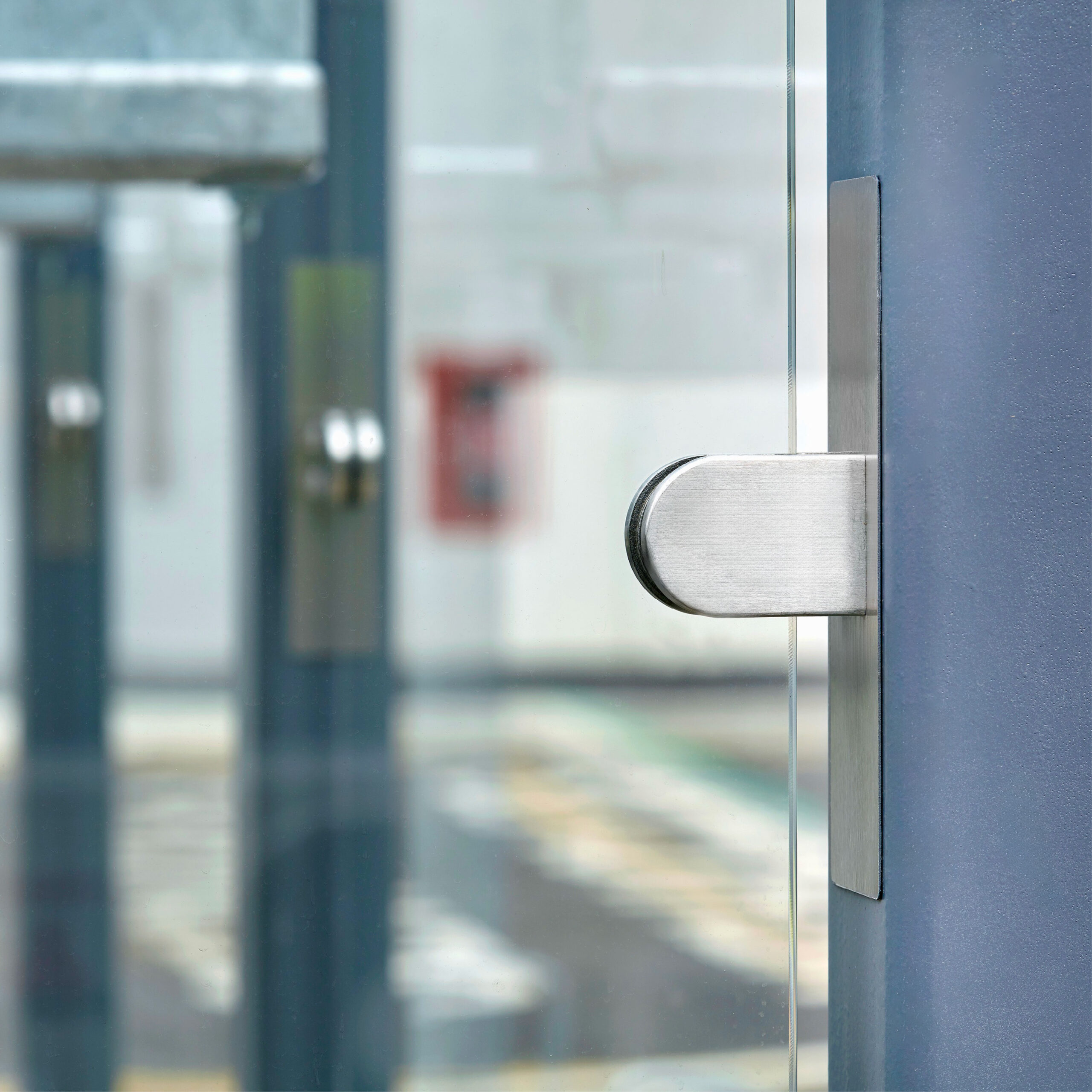 Close-up of a modern, stainless steel door handle on a glass door, showing a blurred urban background with another glass door and a street beyond.