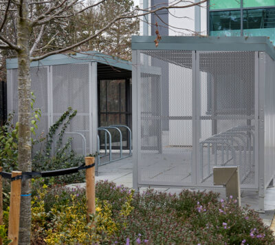a bike rack and metal/mesh bike shelter behind a group of plants