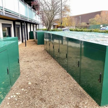 A row of large, green metal storage units, designated as e-bike storage lockers, line the side of a building along a gravel pathway. Another building and trees are visible in the background. The tops of the units are wet, suggesting recent rain.