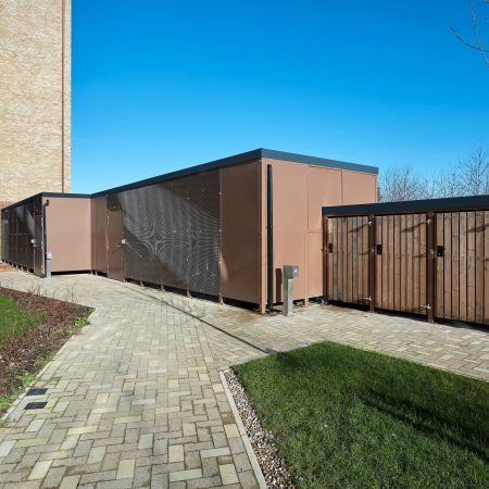 A large brown Carlton Shelter bike enclosure stands prominently beside a brick walkway, inviting visitors to explore its surroundings.