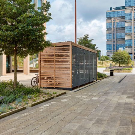 A modern wooden bicycle storage shed with metal elements, located in an urban plaza with paved pathways, landscaping, and tall buildings in the background. a red bicycle is parked next to it.