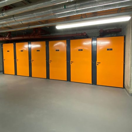 A high-security bike storage area is situated in a corridor with several bright orange doors, numbered, and set in grey frames. The ceiling has exposed pipes and ducts. The floor is clean and smooth, well-lit by overhead fluorescent lights. A directional sign is visible on the left wall.