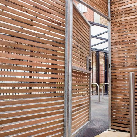 A gate made of horizontal wooden slats framed with metallic rails. The design allows partial visibility through the gaps between the slats. The gate is slightly open, showing part of the structure behind it. The background includes more wooden slats.
